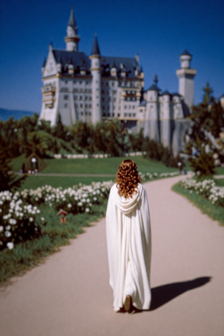 A soft,  dreamy landscape shot of a woman in a white cloak walking away from the camera down a dirt path. In the distance,...