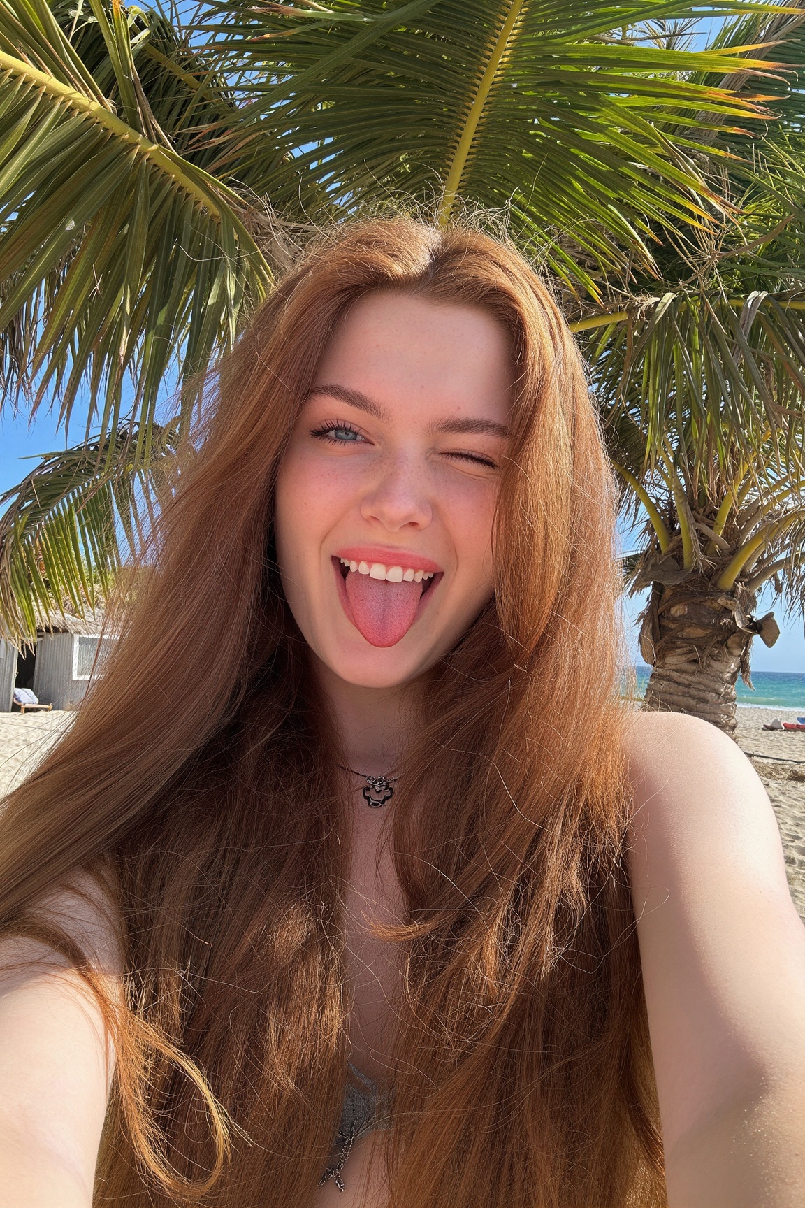 Girl taking a playful beach selfie against a palm tree,  20 years old,  European woman with long wavy auburn hair and frec...