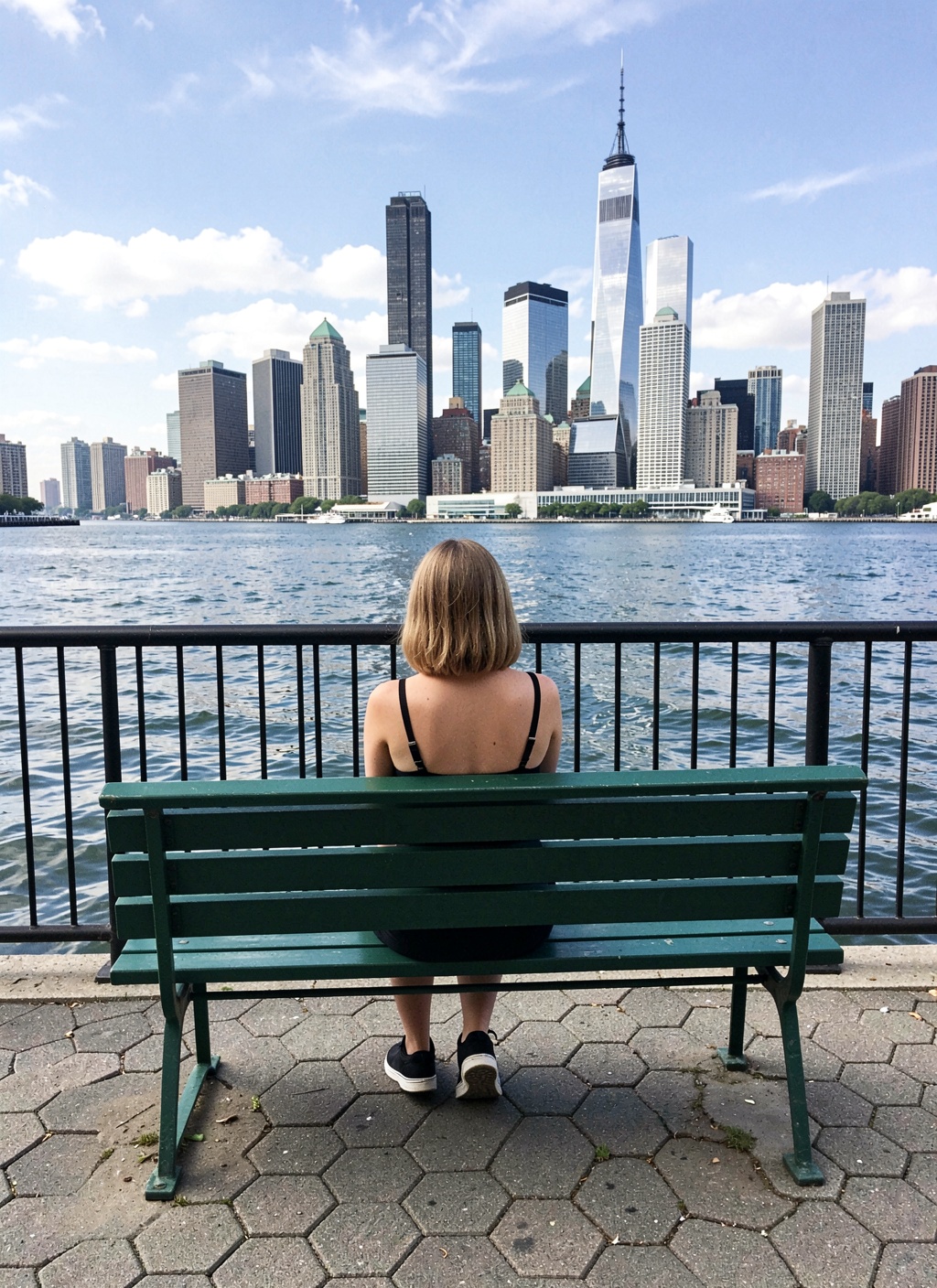 A person with light-brown hair sits on a green metal bench,  facing away from the camera,  looking out over a body of wate...
