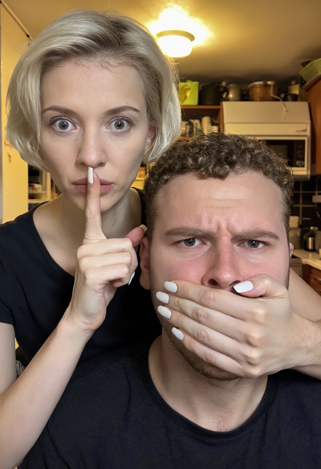 A close-up photograph of a woman covering a man’s mouth and nose with her hand. The woman standing behind the man has soft...