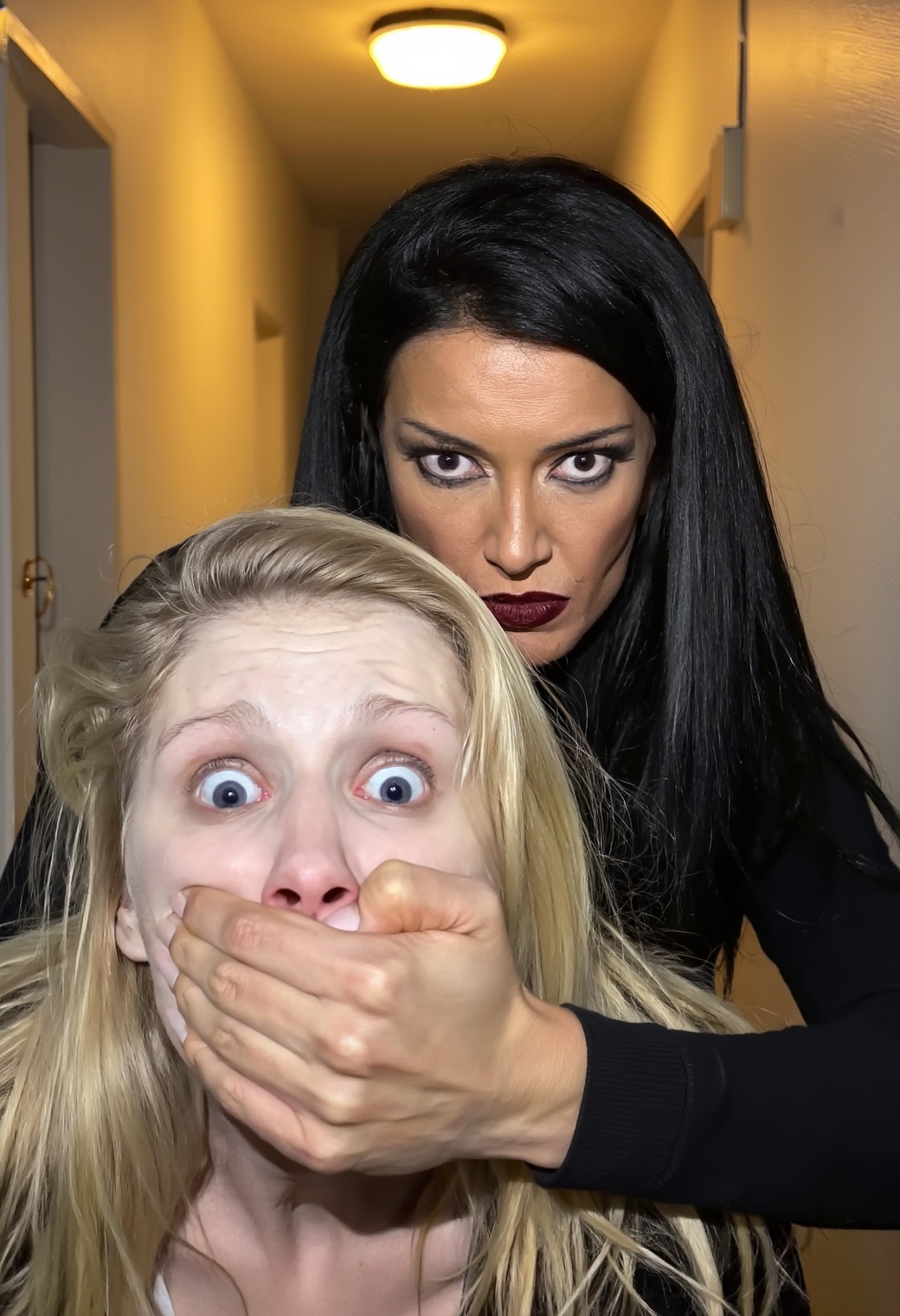 A close-up photograph of a woman covering a another's mouth and nose with her hand. The woman in control has a predatory c...