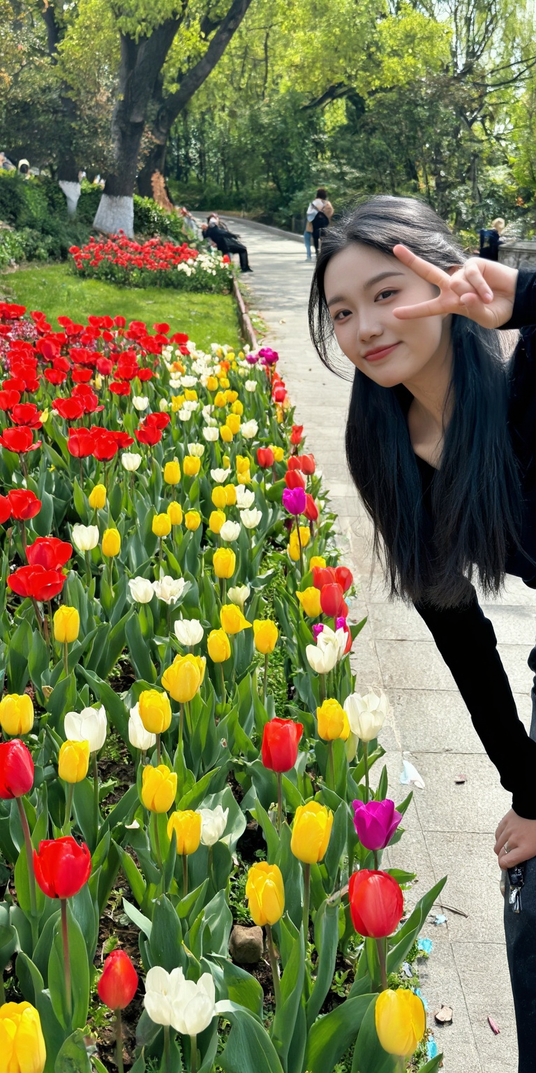a young woman with long black hair,  wearing a black top,  posing playfully near a vibrant flower bed in a park. She is ma...
