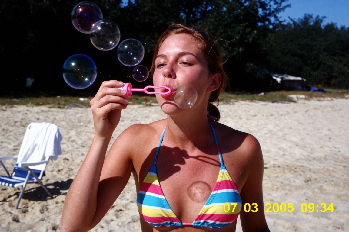 A low-resolution,  candid digital snapshot from 2005 of a young women blowing bubbles on a sandy beach. She has reddish-br...