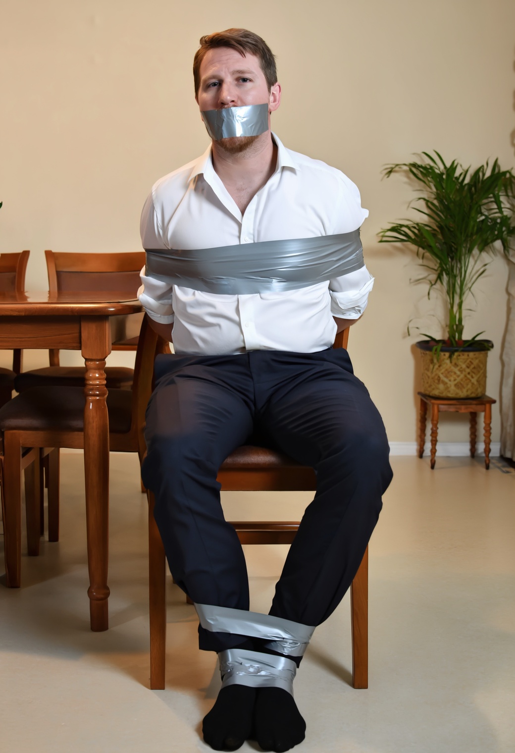 A photo of a man with short brown hair and a light beard sitting on a wooden dining chair. He is dressed in a white dress...