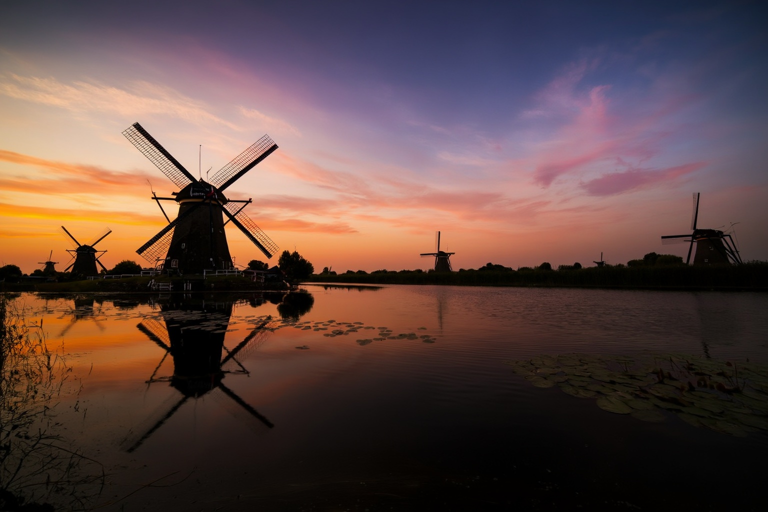 aurelmanea3z photography. A landscape at sunset,  featuring a series of windmills situated on a body of water. The windmil...
