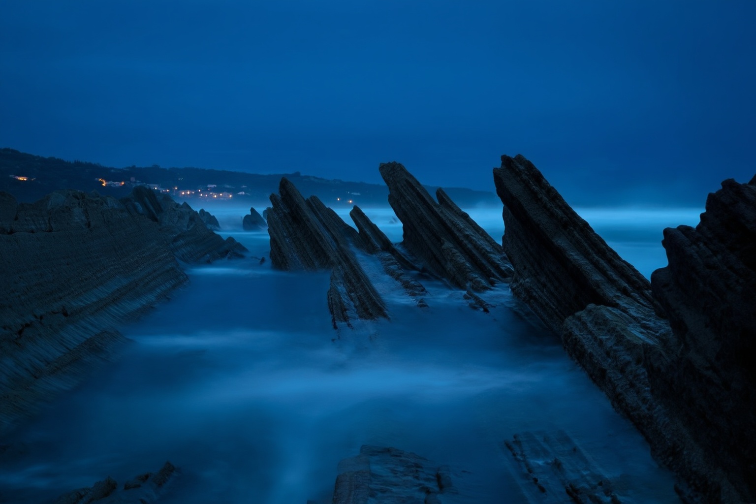 aurelmanea3z photography. A coastal scene at dusk or night,  with a series of jagged rock formations extending into the oc...