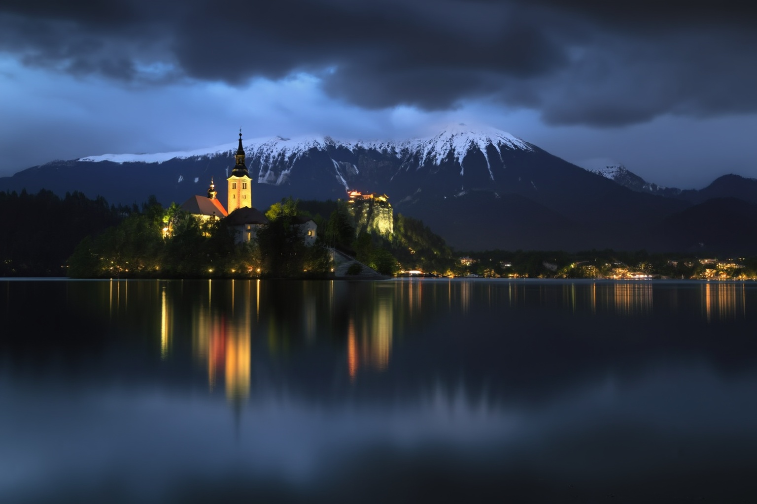 aurelmanea3z photography. A lakeside scene at dusk or nighttime. The foreground features a calm,  reflective lake with a s...