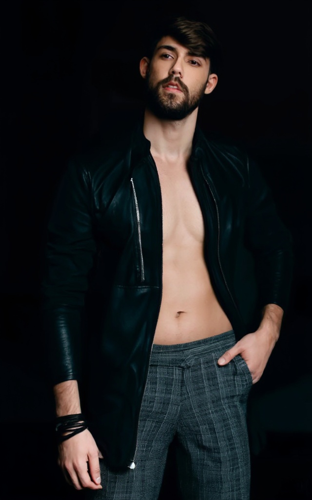 A medium shot of a man stands in a dark room. The man is wearing a black leather jacket. The jacket has a zipper on the ri...