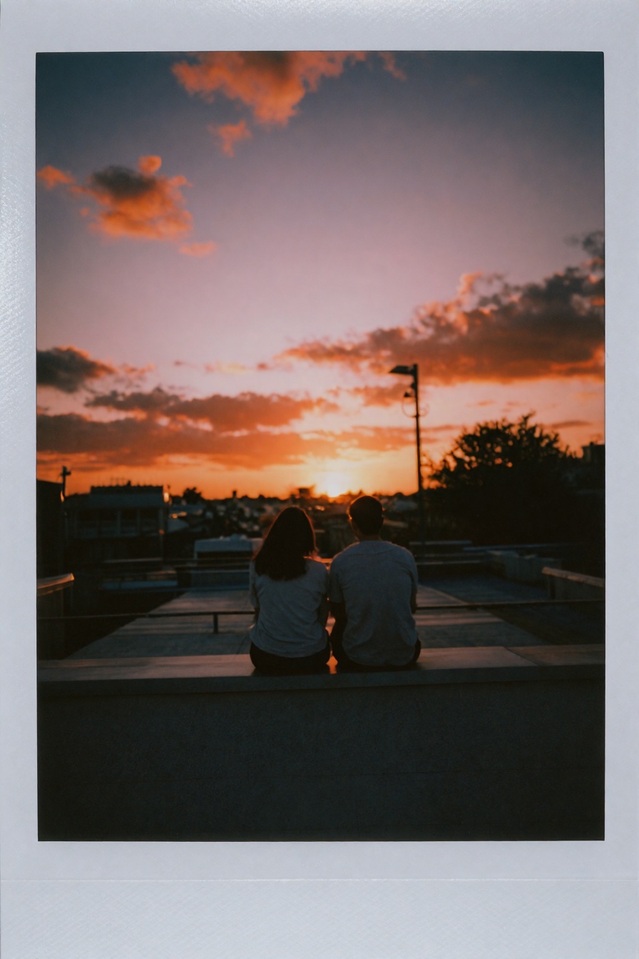 Candid Instax Photo,  The image is a photograph of two people sitting on a rooftop facing away from the camera. The sky is...