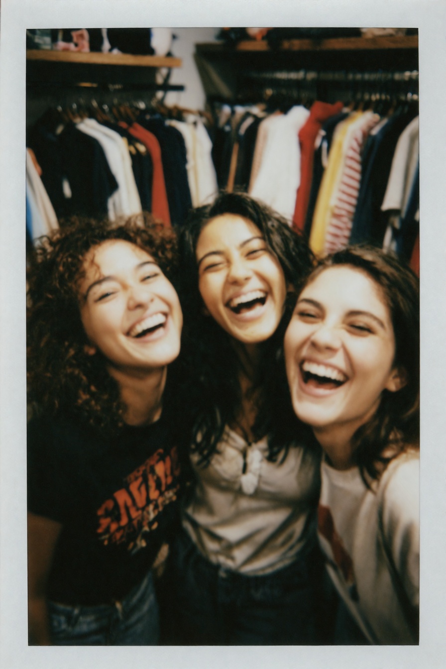 Candid Instax Photo,  The image is a polaroid photograph of three young women laughing together. They are standing in fron...