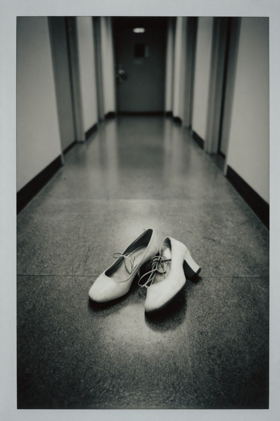 Candid Instax Photo,  The image shows a pair of white high-heeled shoes lying on the floor of a long hallway. The hallway...
