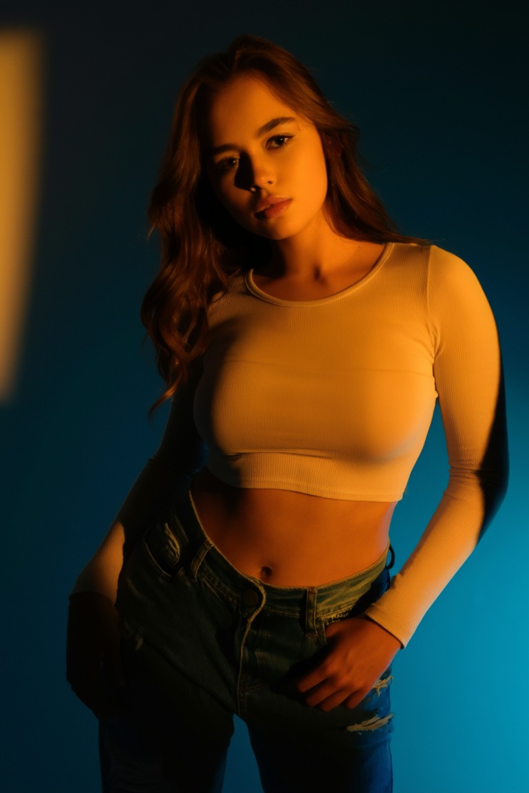 A young woman stands in a tight medium shot against a smooth gradient wall — warm golden light from the left casts a soft...