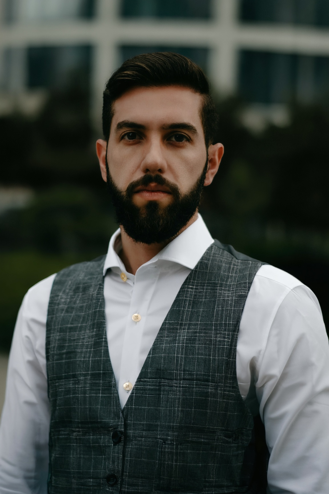 A man with dark,  neatly combed hair and a full beard stands against a modern building. He wears a white dress shirt and a...