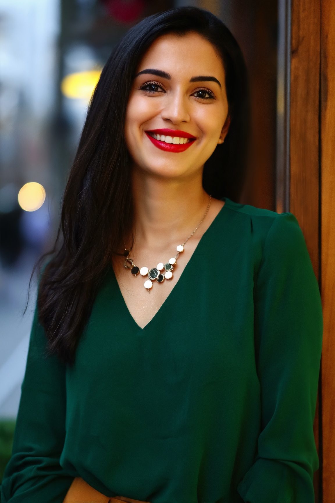A woman with long dark hair smiles warmly,  dressed in a simple dark green blouse. Her bright red lipstick stands out agai...