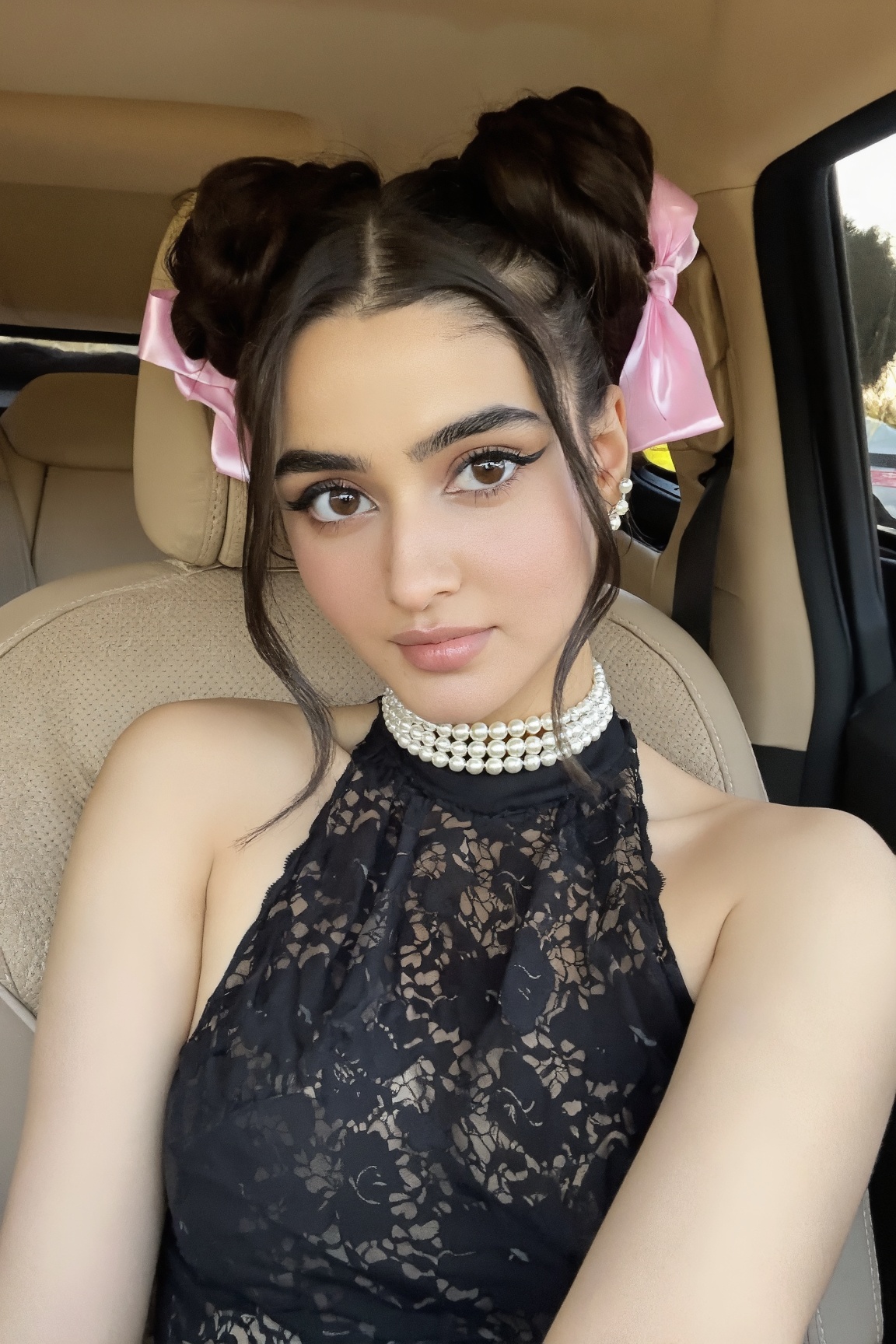 A young woman sits in a car’s front seat,  hair styled in twin buns with pink ribbons. She wears a black lace halter top w...