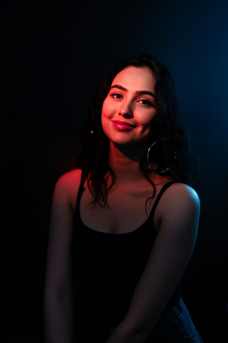 A young woman with long dark wavy hair (appears lightly damp) sits against a dark muted backdrop. She wears a simple black...