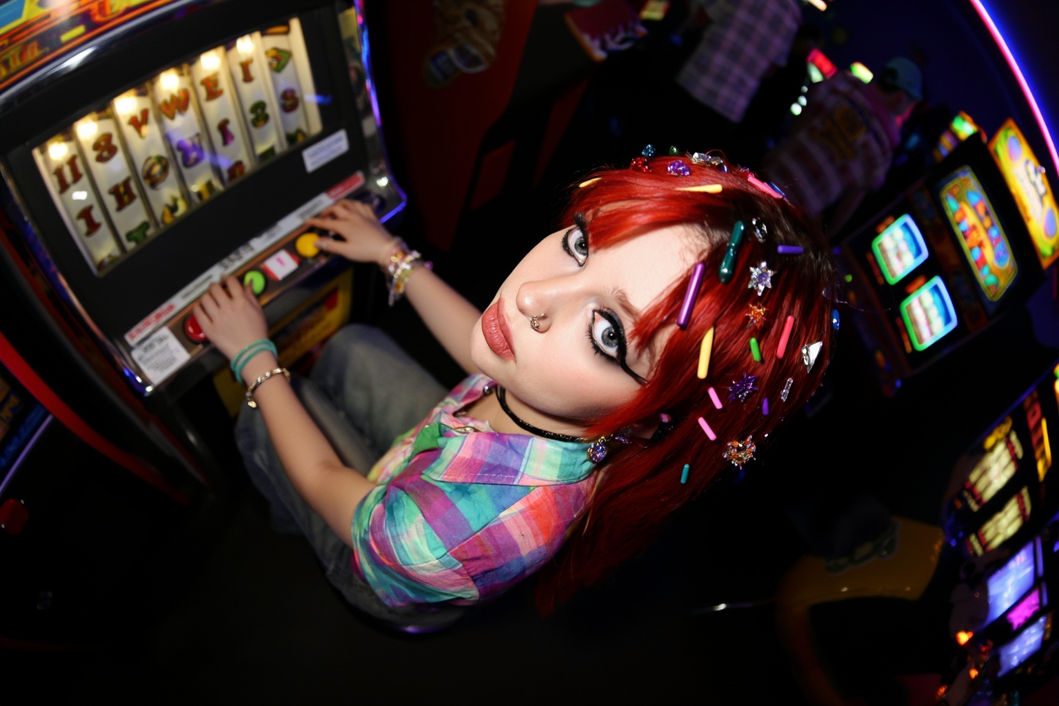 A high-angle fisheye shot looking down at a girl with bright red hair decorated with colorful stick-on gems. She's sitting...