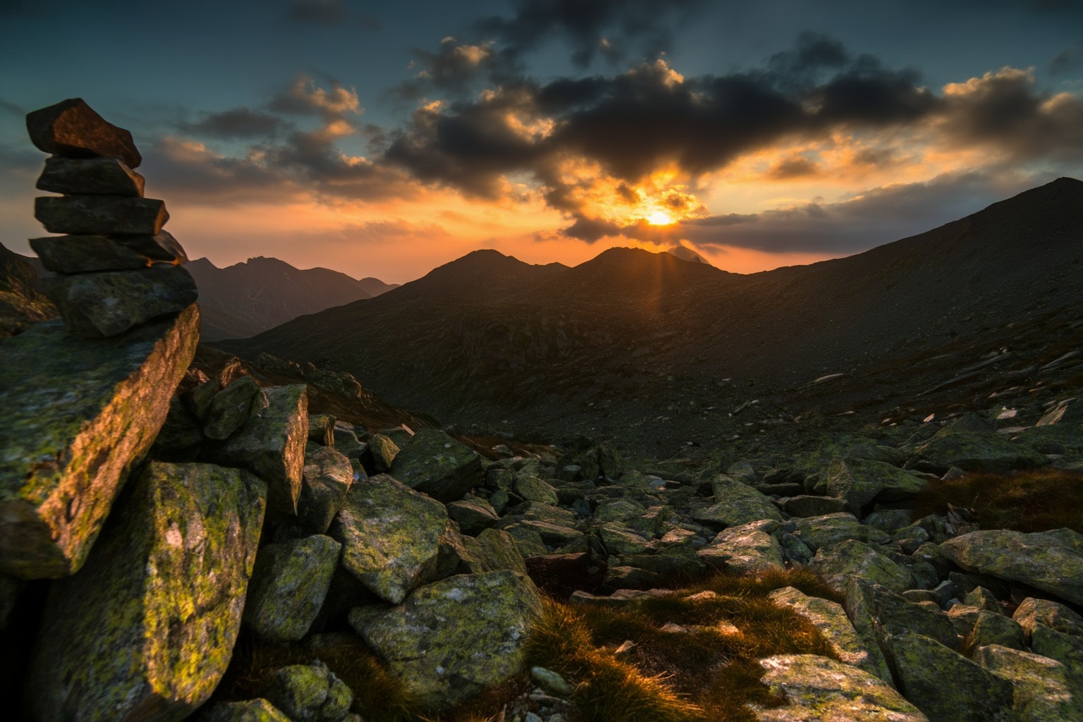 aurelmanea5z photography. Photograph of a rugged mountain landscape at sunset. The sun is partially obscured by dark,  clo...