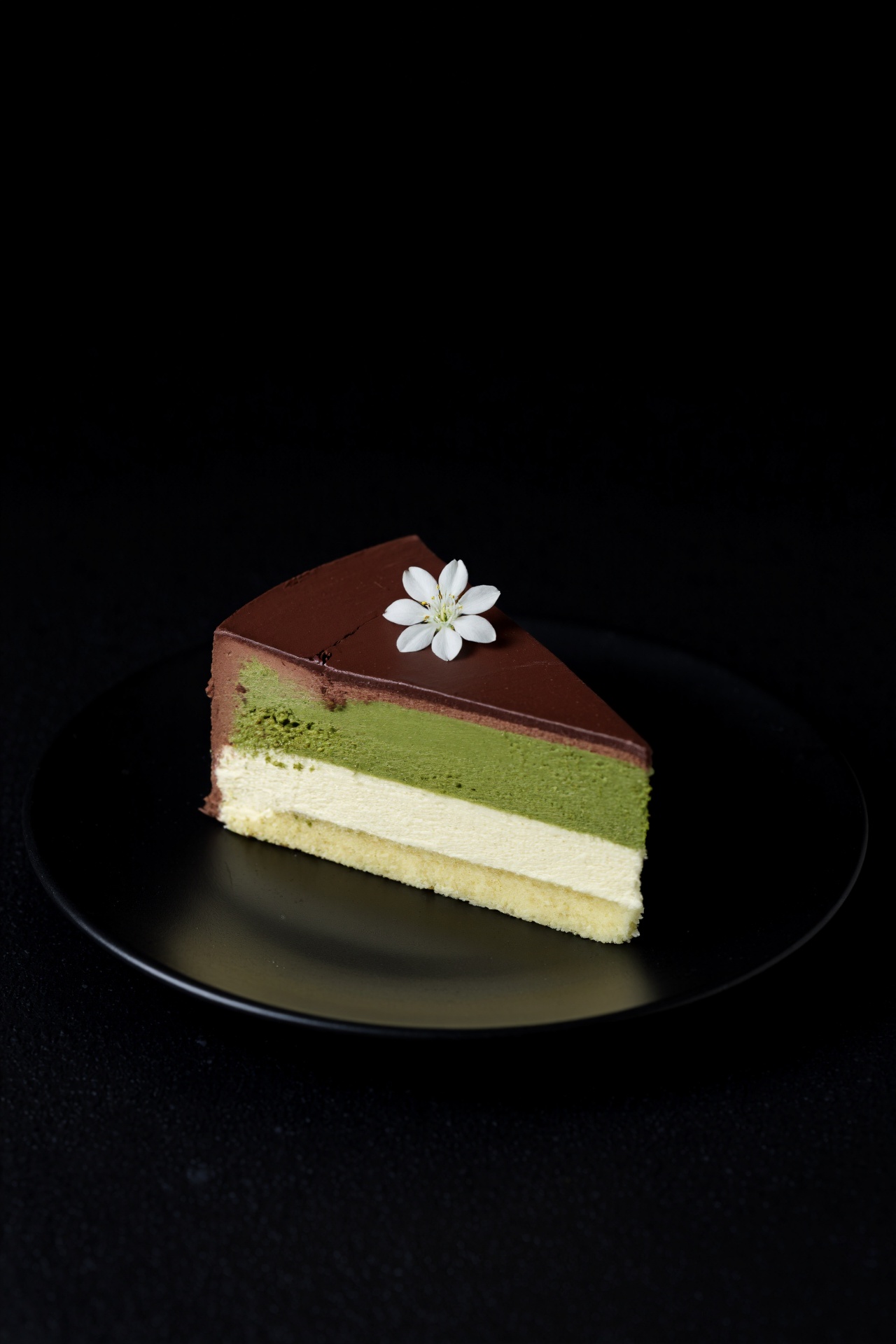 a slice of cake with chocolate,  green tea,  and vanilla cream on a black plate. a small white flower is placed in the cen...