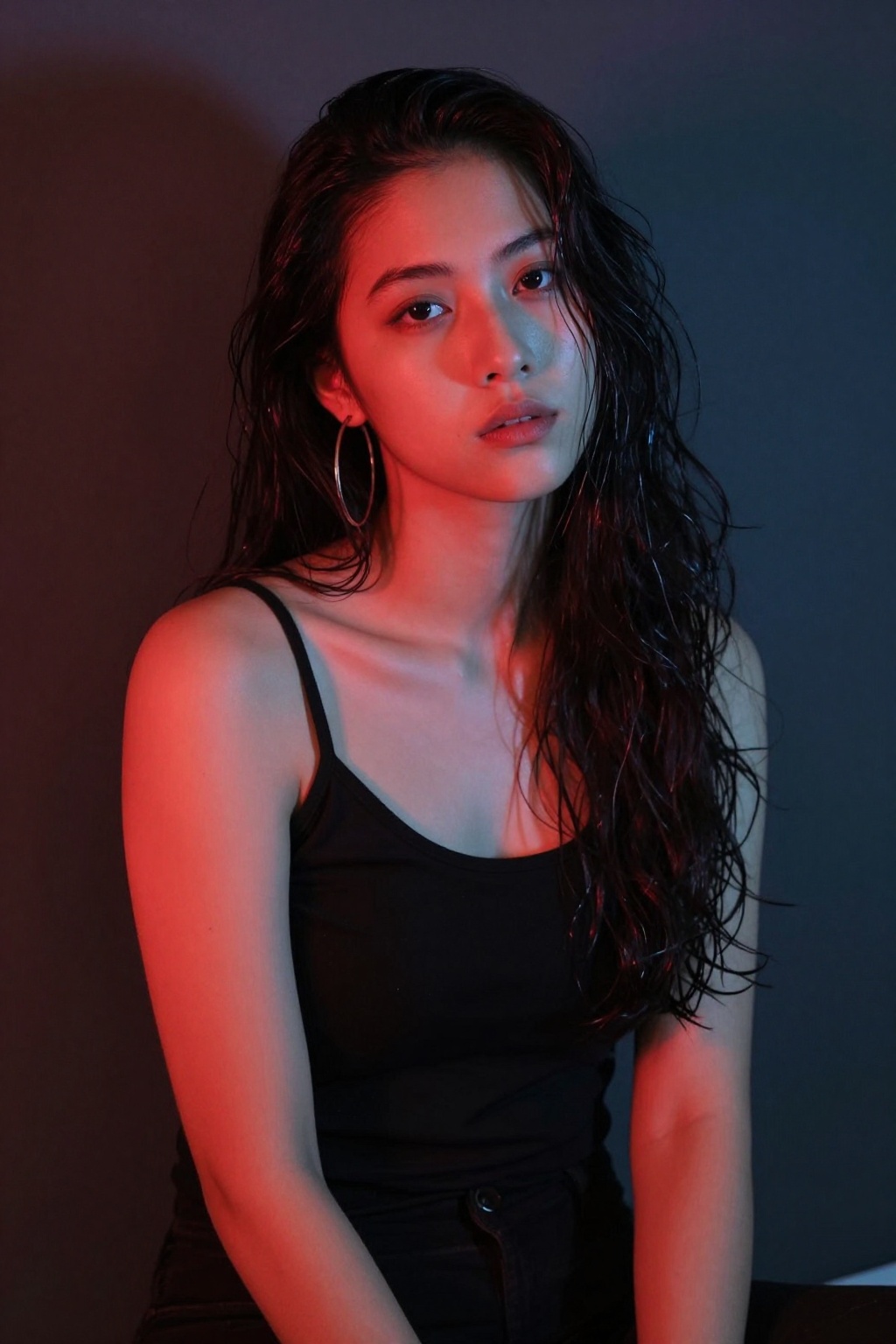 A young woman with long dark wavy hair (appears lightly damp) sits against a dark muted backdrop. She wears a simple black...