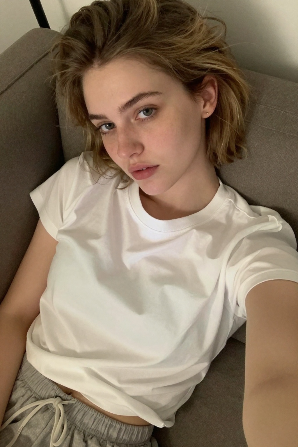 Low-angle casual selfie of a 20-year-old woman with short,  tousled dark blonde hair tucked behind one ear. Her skin is ve...