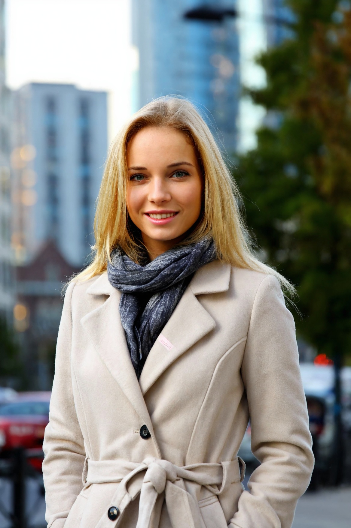 A candid photo of the young woD4n1 with blonde hair. She is wearing a beige coat and a scarf. The background is blurred an...
