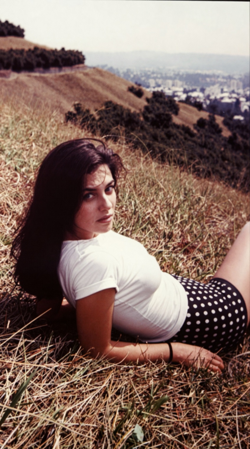 "A vintage 35mm film photograph of a young woman with long dark hair lying on her stomach on a dry,  grassy hillside,  loo...
