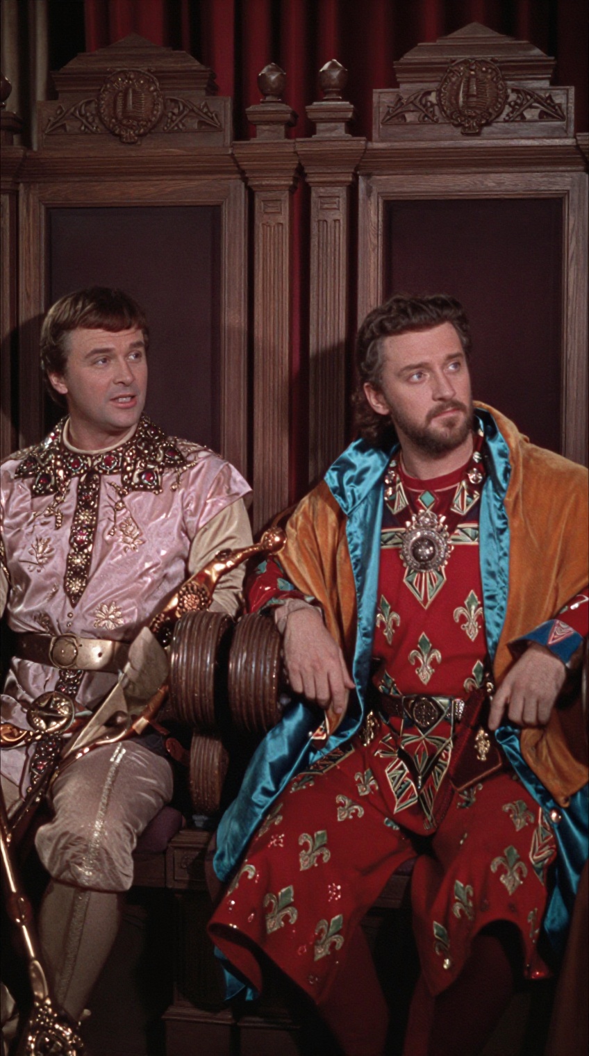 Two men in ornate medieval-style regalia sit side by side on elaborately carved wooden thrones against a deep red curtain...