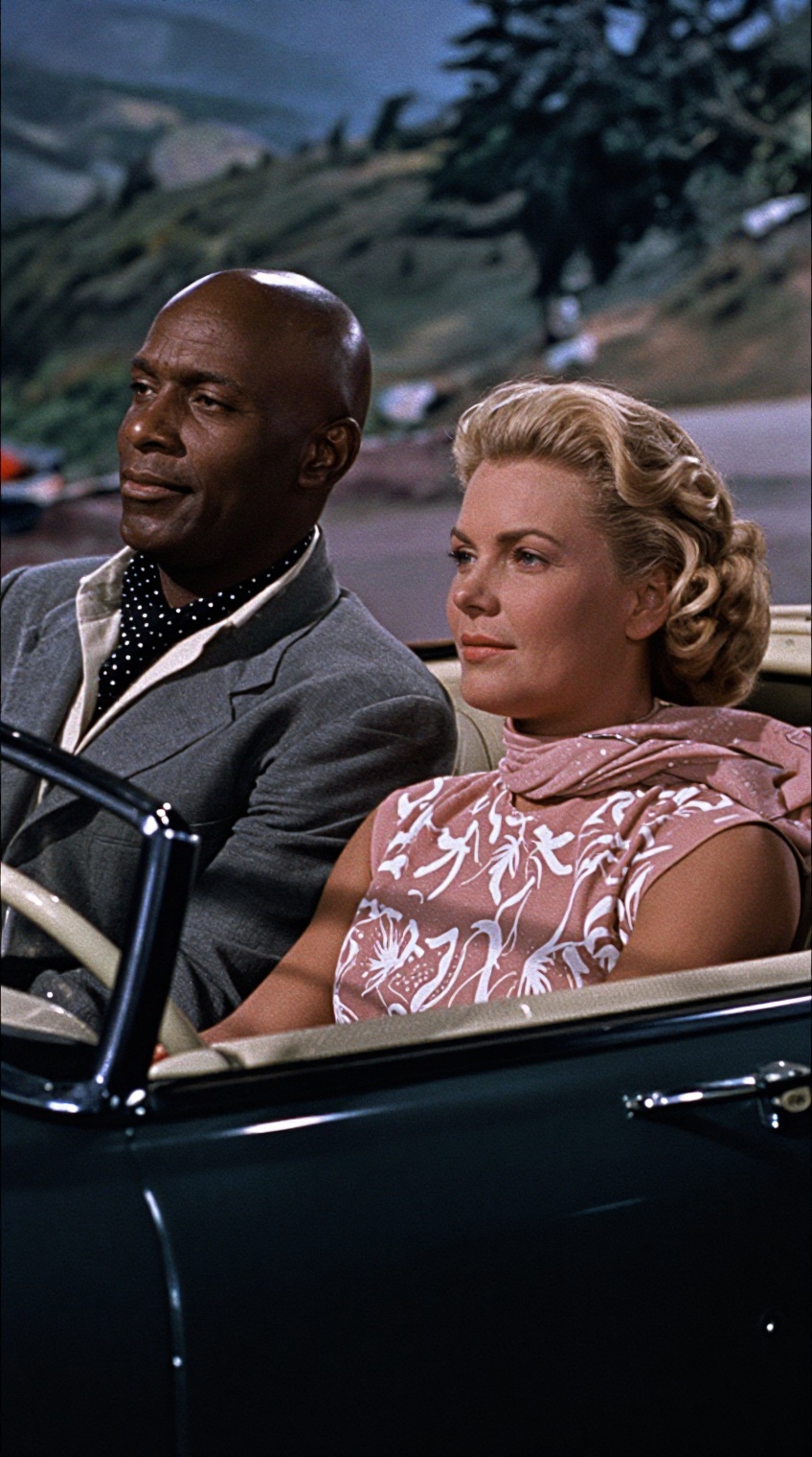 A bald,  black african man in a gray suit with a black polka-dot scarf sits in the passenger seat of a dark convertible,...