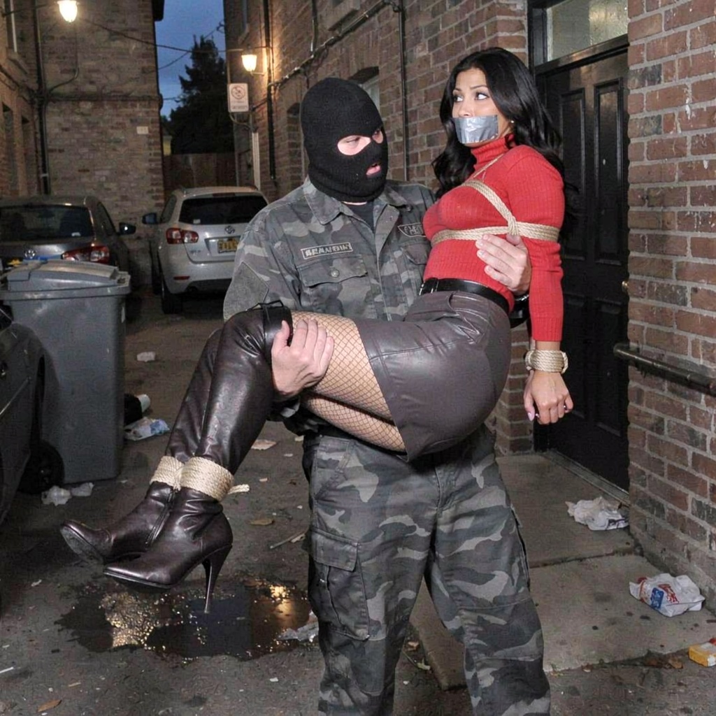A caucasian man wearing a full face balaclava that leaves visible his eyes and mouth carries a bound woman in a princess c...