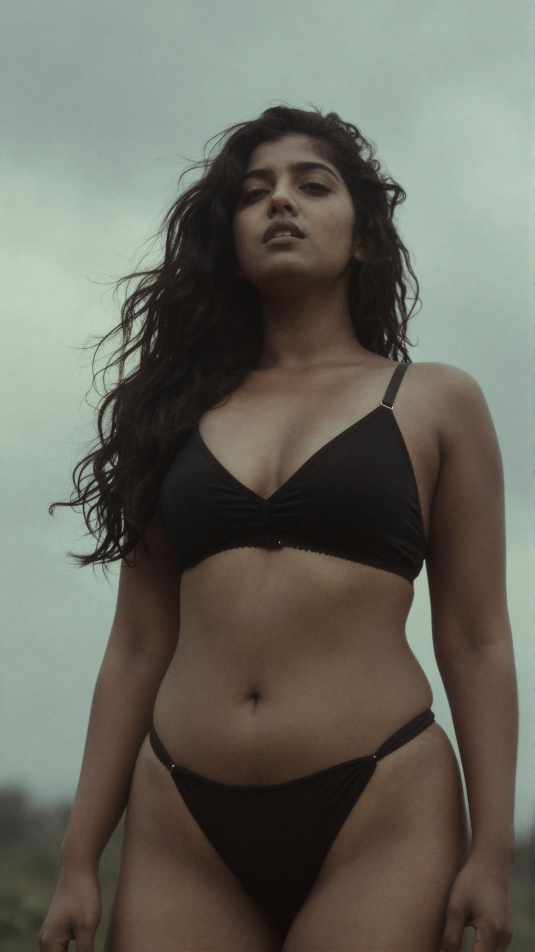 A hauntingly beautiful photograph of a young woman with long,  tousled dark hair,  wearing a black black bra and lowest wa...
