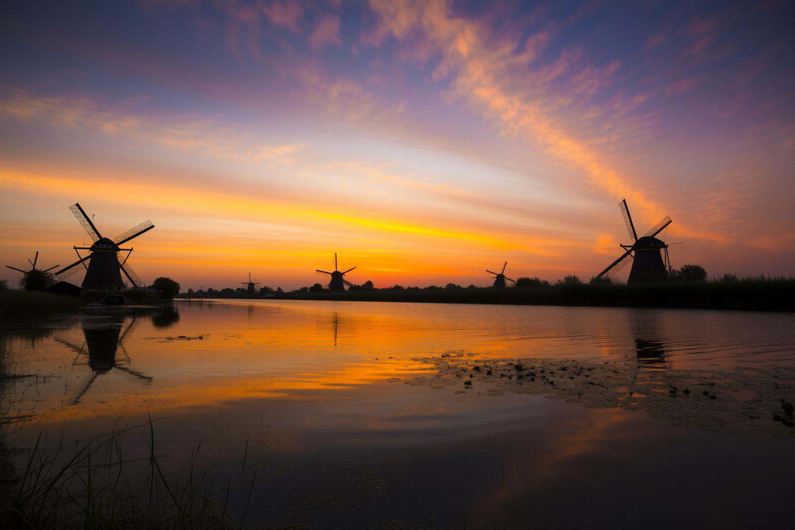 aurelmanea2q photography.A landscape at sunset,  featuring a series of windmills situated on a body of water. The windmill...