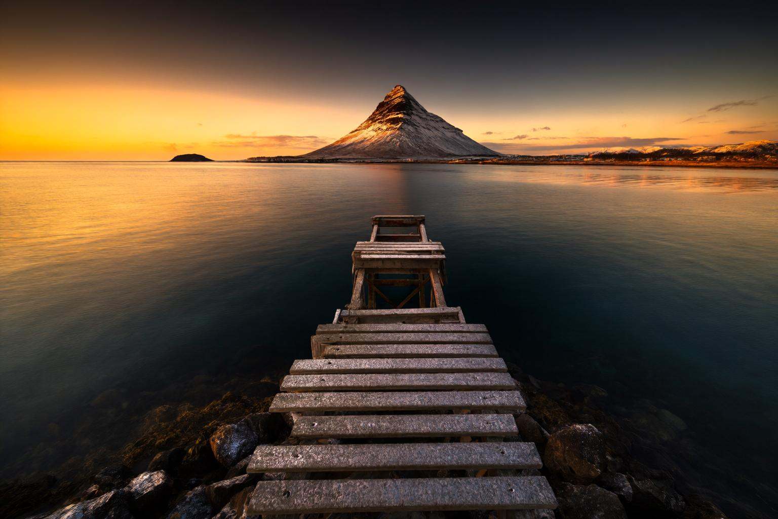 aurelmanea2q photography. A seascape at sunset,  with a wooden dock extending into the calm water. The dock leads to a sma...