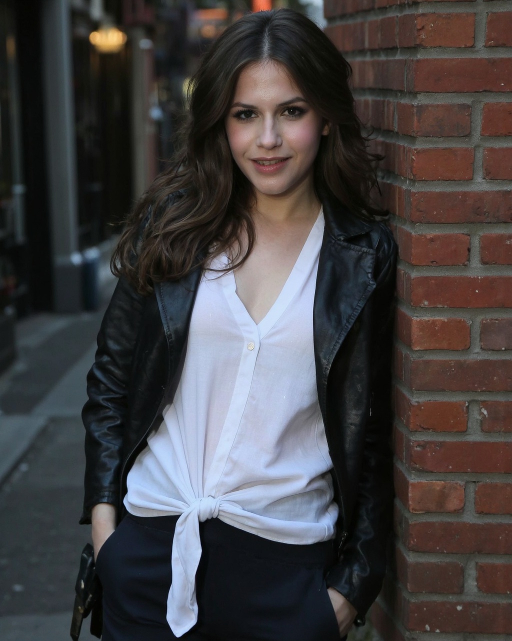 A portrait of erin_sanders leaning against a brick wall. She wears a white shirt tied at the front,  a black leather jacke...