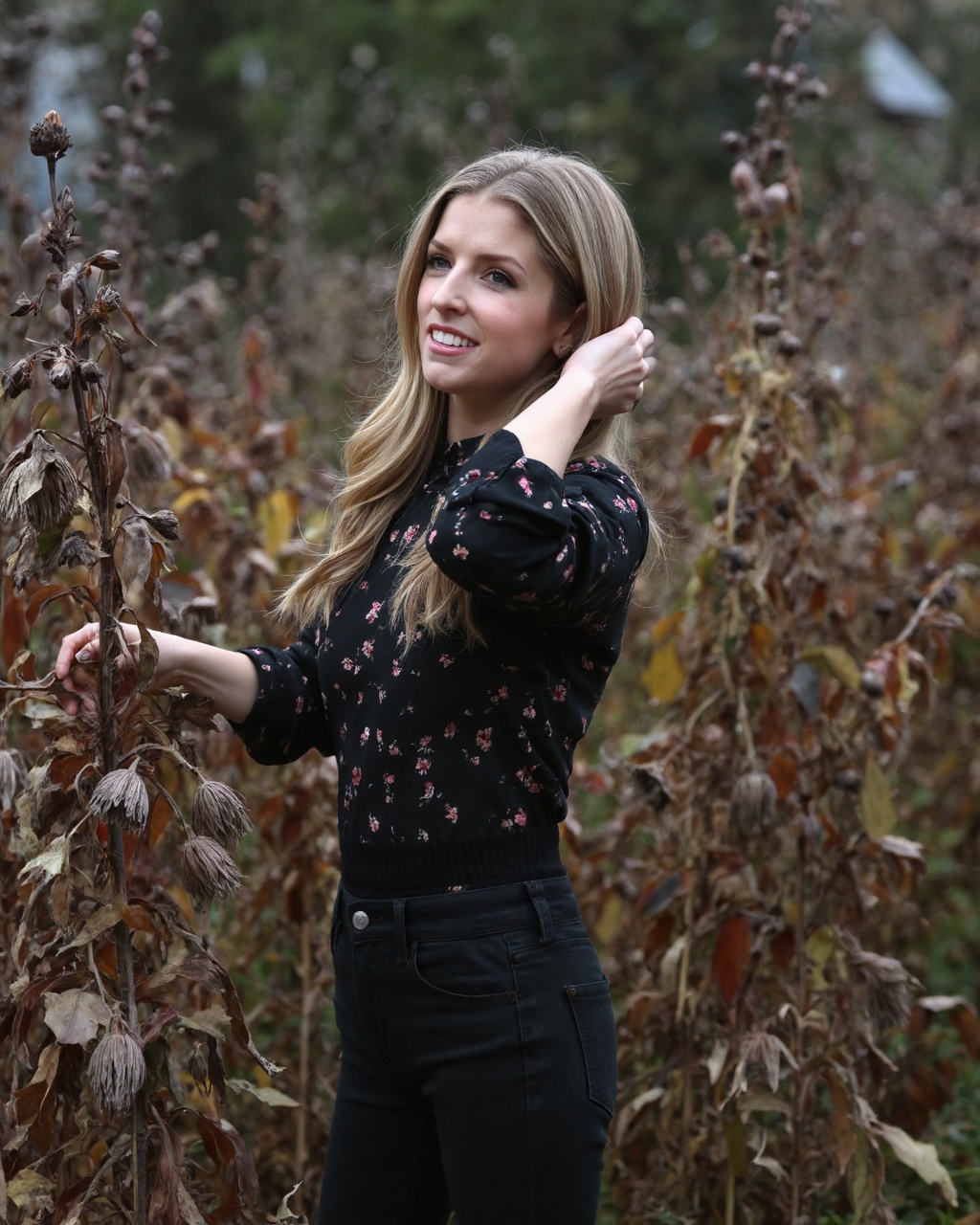 Anna_kendrick standing amidst a dense thicket of tall,  dried wildflowers in a natural,  forested setting. The woman is po...