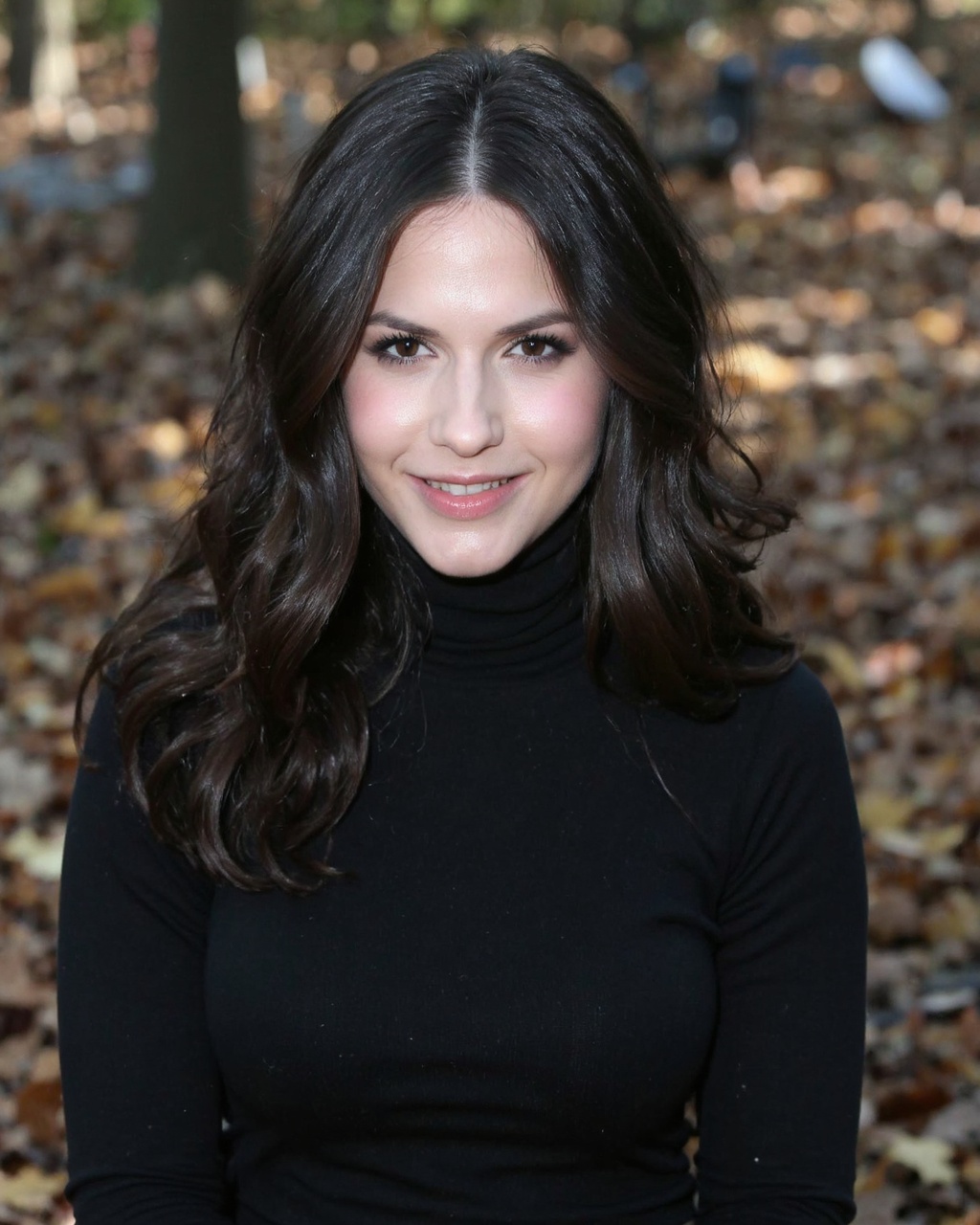 A portrait of erin_sanders outdoors. She has long,  dark hair and wears a black turtleneck sweater. Her gaze is directly a...