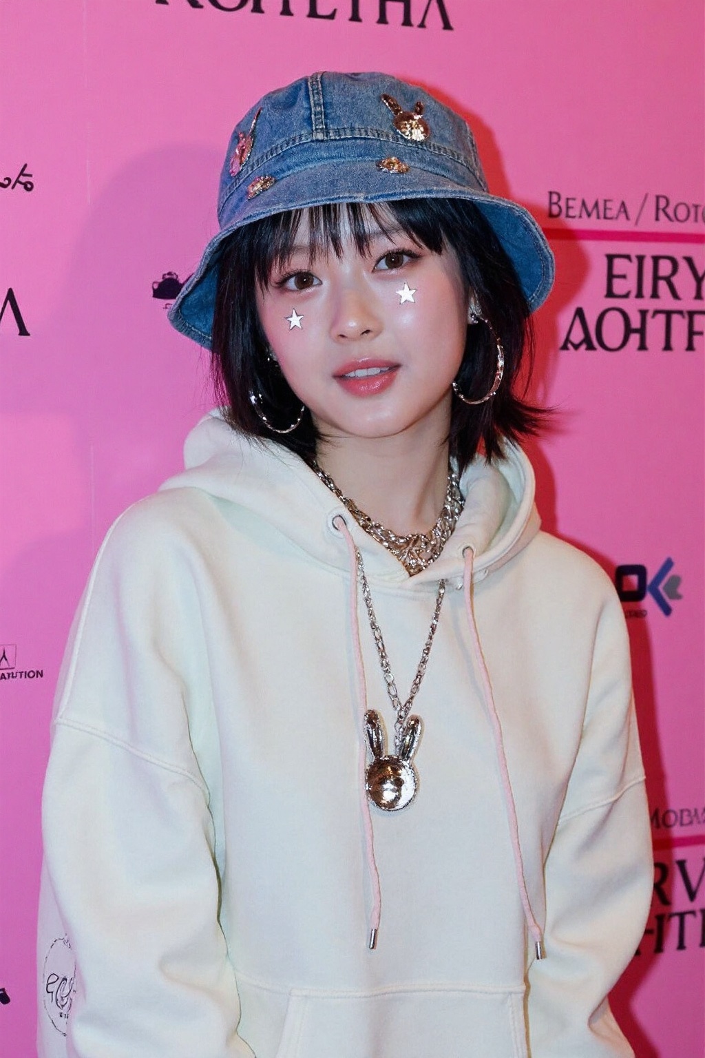The image shows a petite young asian woman standing in front of a pink background with text on it,  possibly part of a pos...