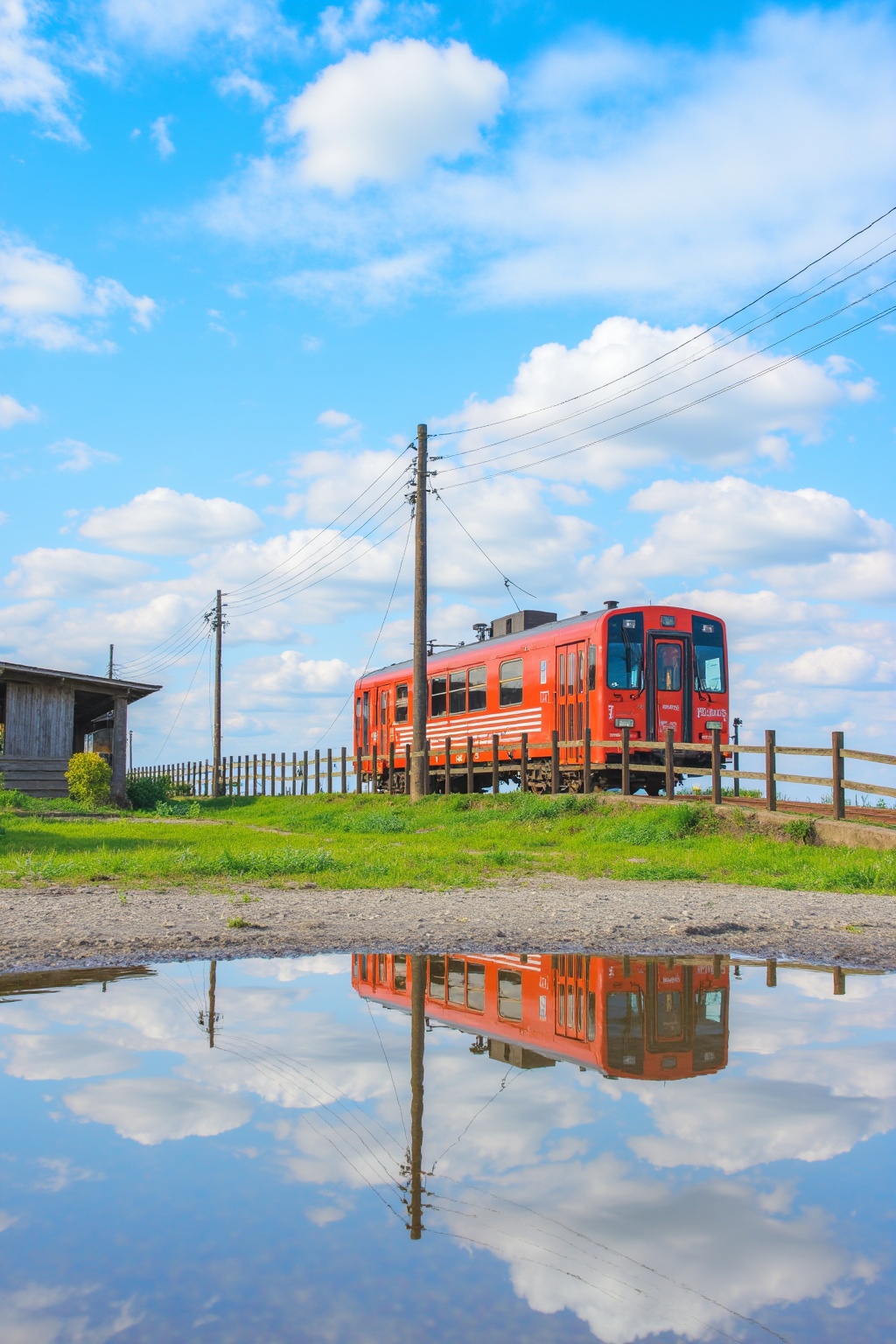 a red train traveling down tracks next to a puddle of water. The train is surrounded by a fence, grass, poles, wires, and a shed. The sky in the background is filled with clouds.