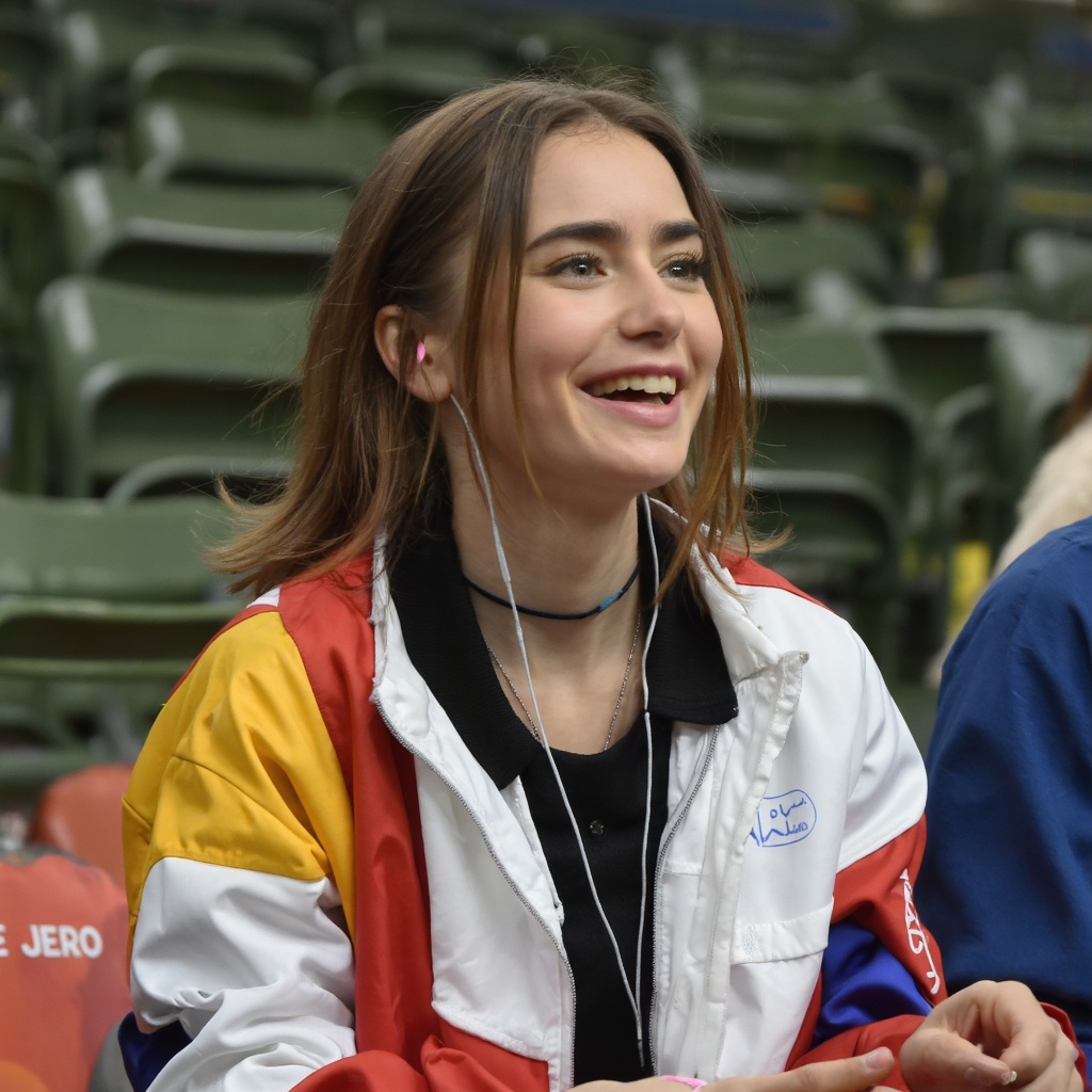 A portrait of lily_collins seated in an indoor stadium. She wears a white jacket with red,  yellow,  and blue accents on T...