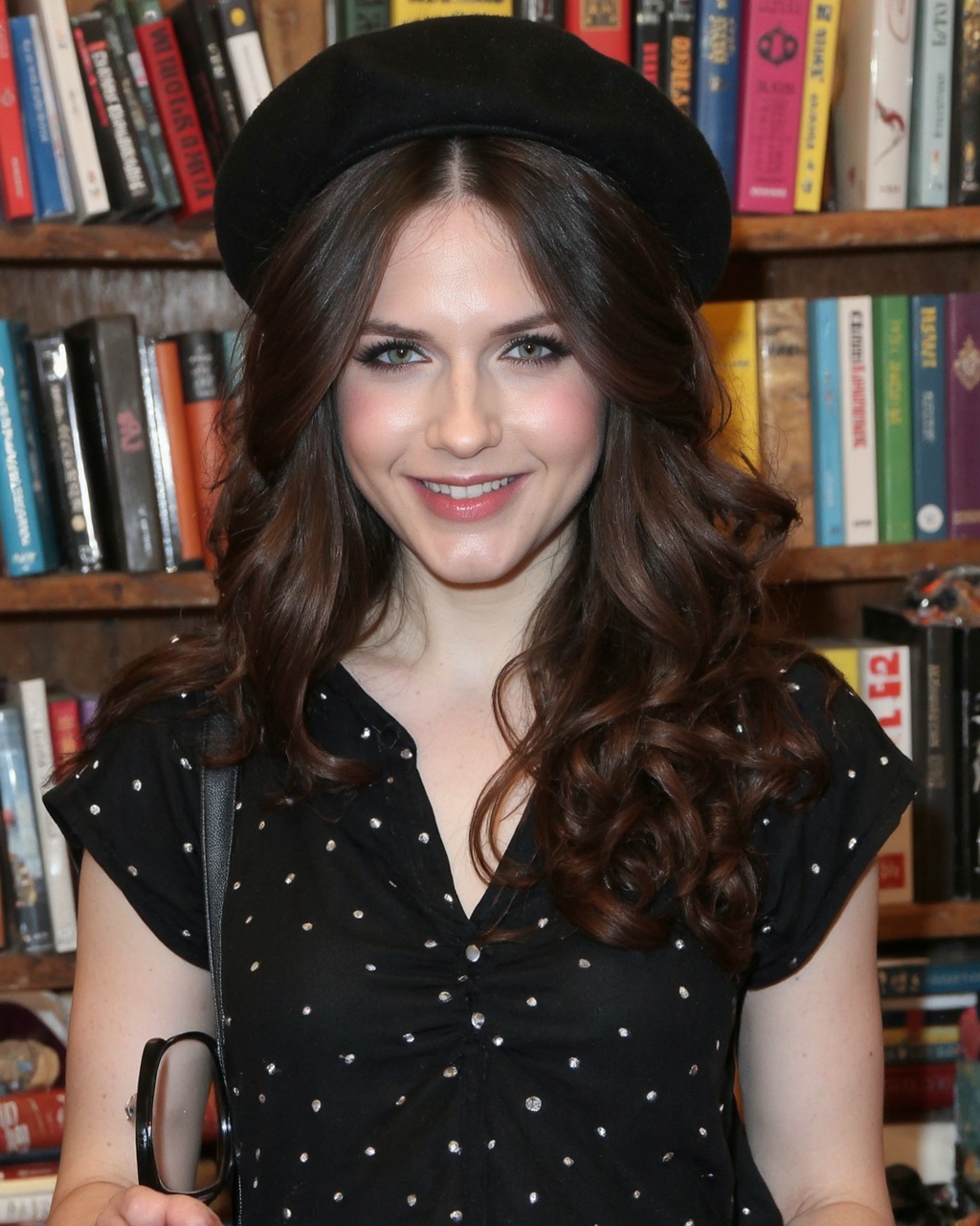 A portrait of erin_sanders in a bookstore. She wears a black beret and a black blouse with white polka dots. Her long brow...