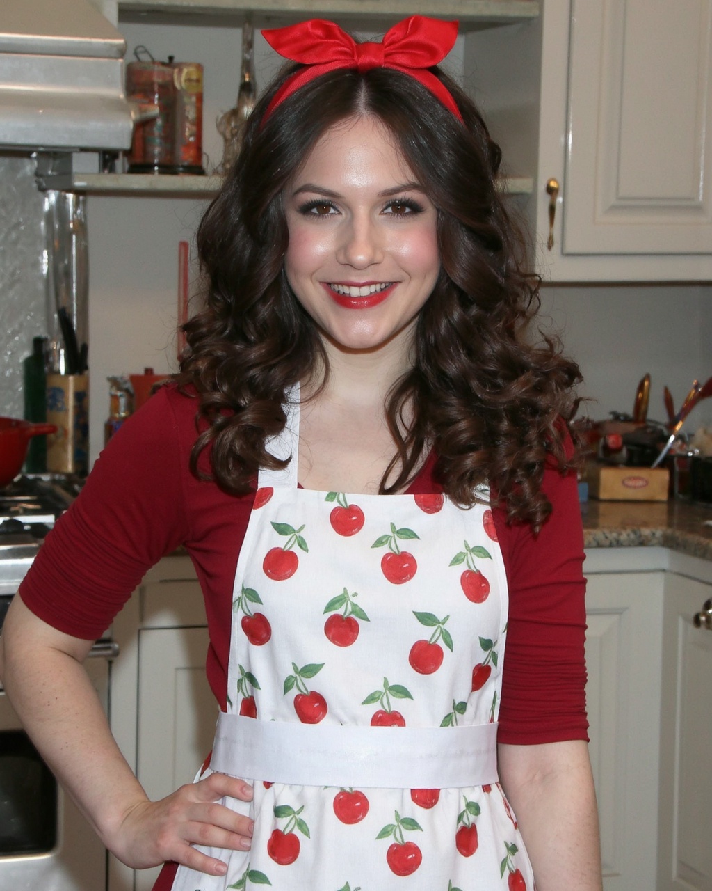 A portrait of erin_sanders in a vintage kitchen. She wears a white apron with red cherries and white polka dots,  and a re...