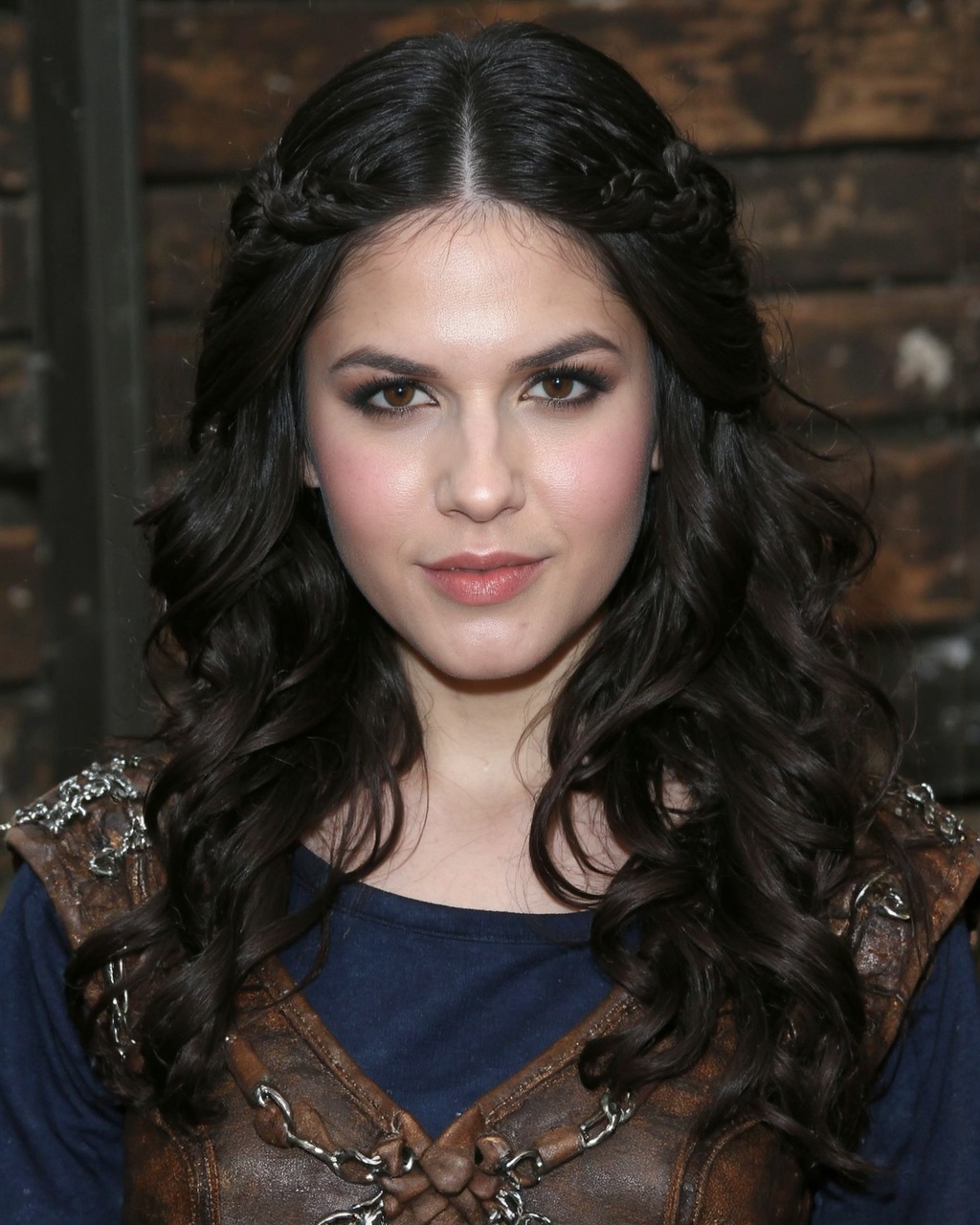 A portrait of erin_sanders with long,  wavy black hair styled in a braided updo,  set against a rustic wooden backdrop. Sh...