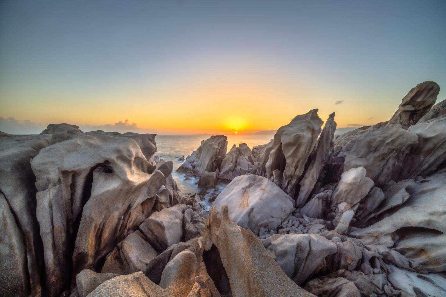 aurelmanea2q photography. A stunning coastal scene at sunset. The foreground features rugged,  eroded rock formations with...