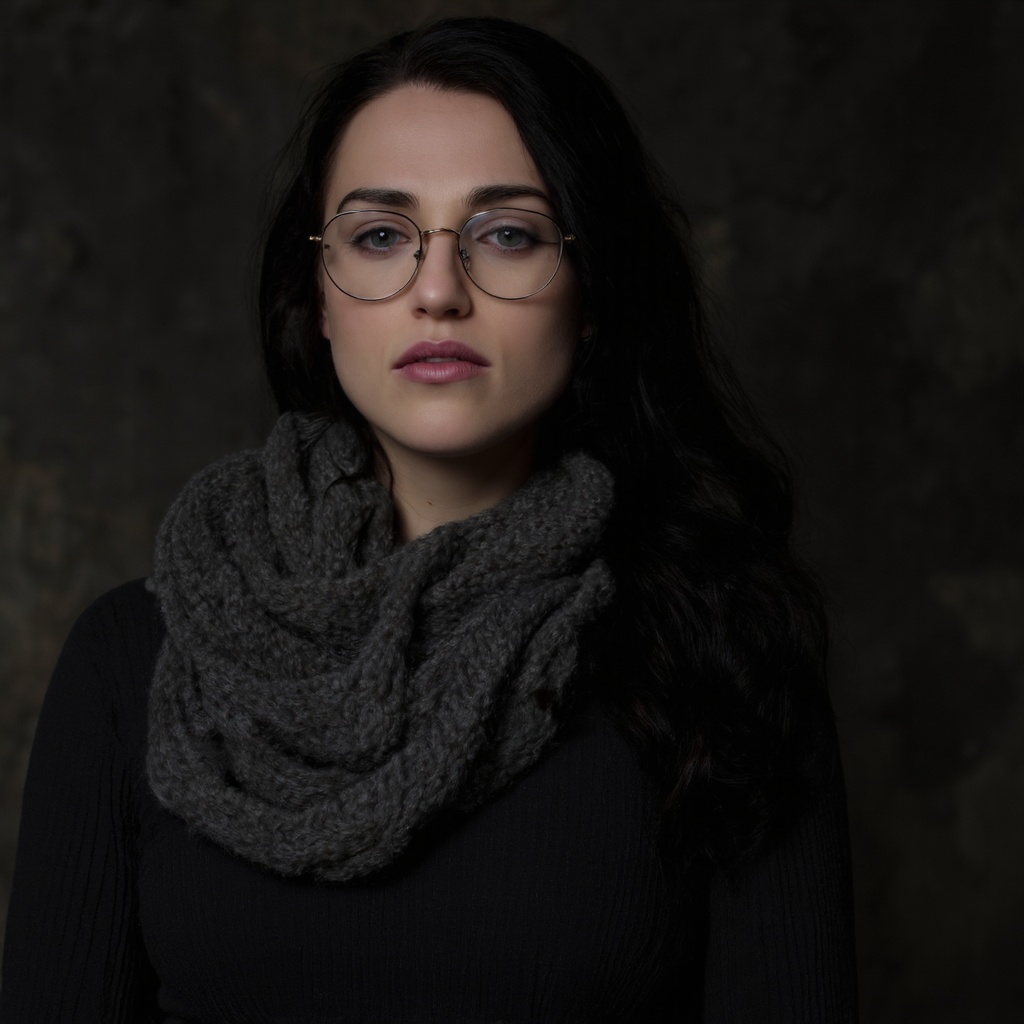 A portrait of katie_mcgrath against a dark,  textured background. She wears round,  gold-framed glasses and a textured gra...