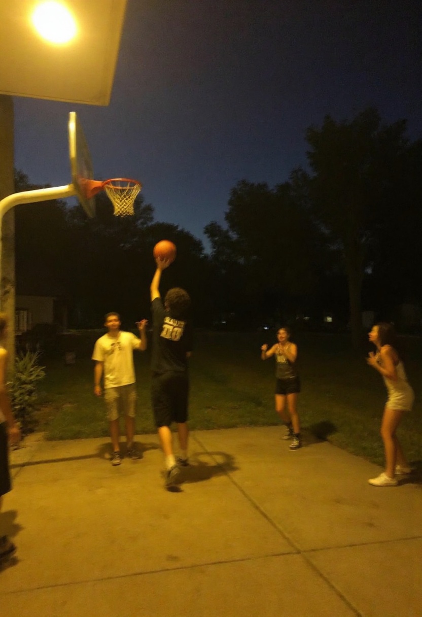 This photograph captures A group of friends playing a casual game of basketball in someone’s driveway at dusk. One friend...