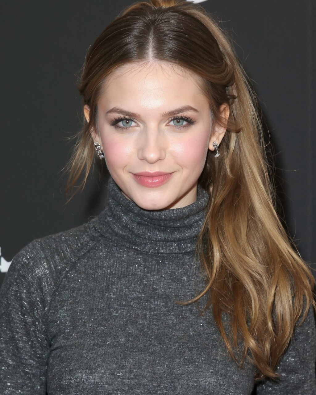 A portrait of erin_sanders with long,  flowing blonde hair. She wears a textured gray turtleneck sweater and has a neutral...