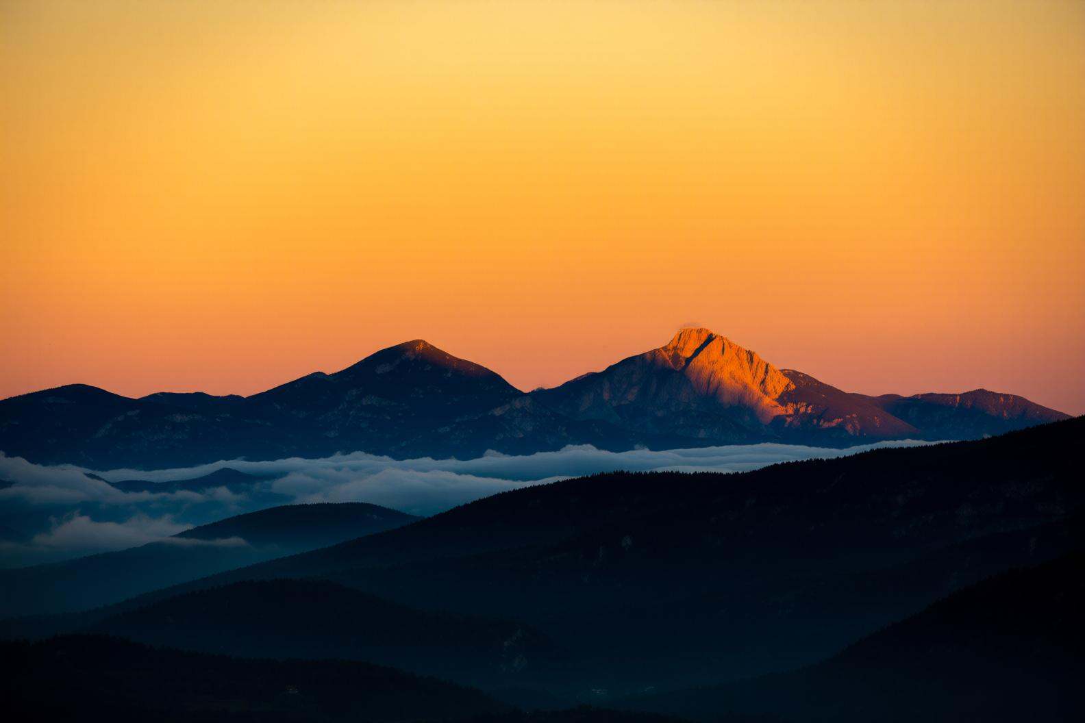 aurelmanea2q photography.A mountain landscape at sunrise. The sky is a gradient of warm orange and yellow hues,  creating...