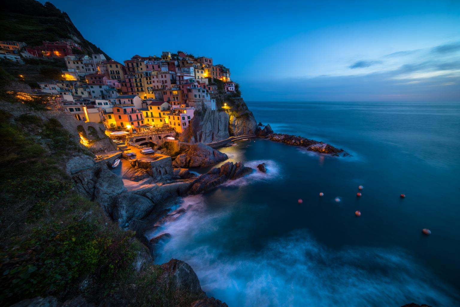 aurelmanea2q photography. A picturesque coastal village at dusk,  with colorful buildings perched on a steep cliffside ove...