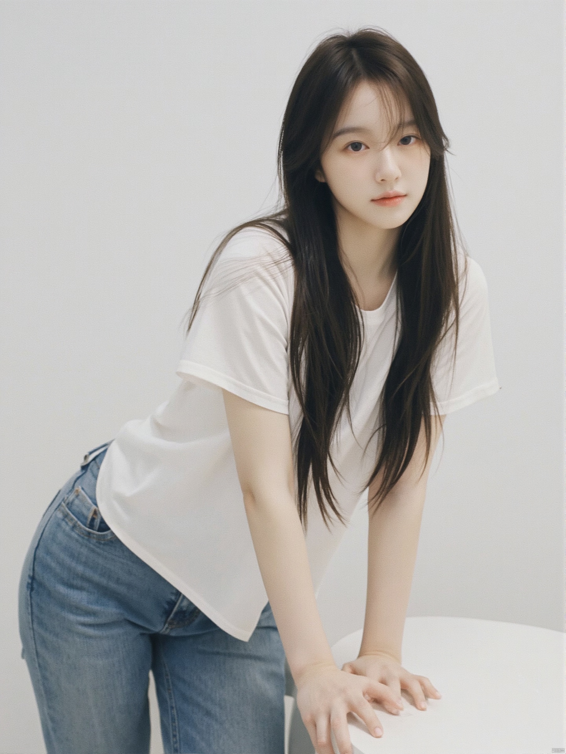 a young Asian woman with long dark brown hair, wearing a white short-sleeved t-shirt and jeans, is leaning on a white table. Her left hand is resting on the table, adding a touch of touch to the scene. The backdrop is a stark white wall, creating a stark contrast to the woman's white shirt.