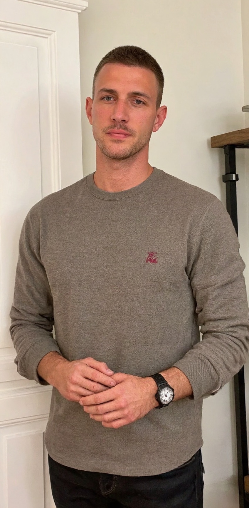 a medium-sized man stands in front of a white door. He is dressed in a long-sleeved gray button-down shirt,  adorned with...